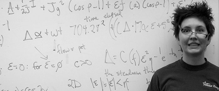 Closeup of person with bushy hair and titled glasses making goofy expression while standing in front of a whiteboard full of equations. 