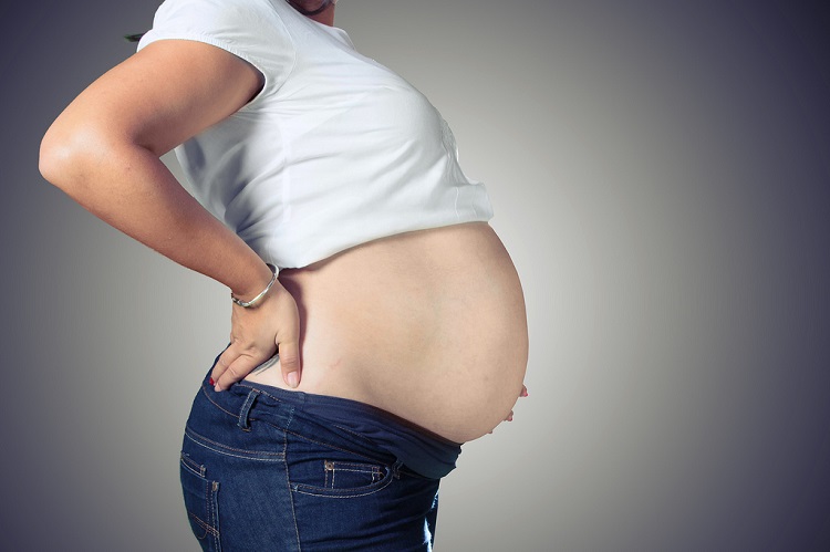 Late-stage pregnant woman, head cropped, arms akimbo, wearing tee shirt and jeans, bare belly protruding. 