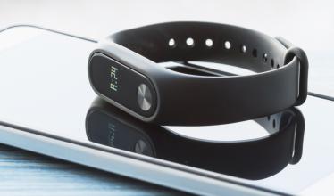 A fitness tracker watch with a black band and narrow, elongated screen sits atop a smartphone, taking up nearly the entire frame.