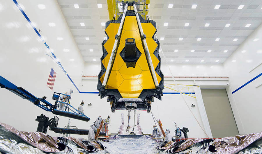 A piece of James Webb Space Telescope hangs from the ceiling as engineers below it work to assemble it.