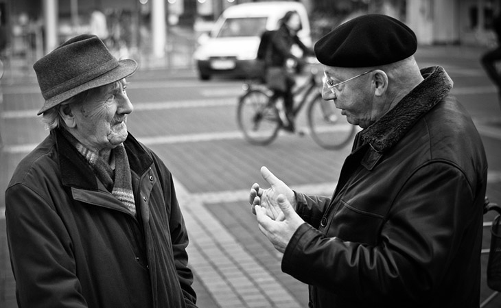 Two older men talking on a city street with a cyclist in the background. 