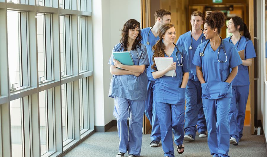 Several medical students wearing blue scrubs walk in a hallway.