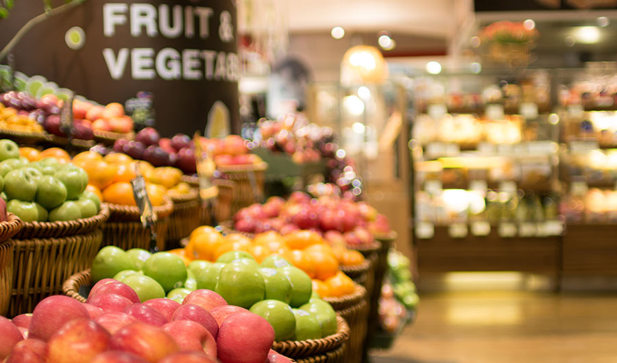 Image shows a grocery store, with apples and other fruit in the foreground with more of the market in the background and out of focus.