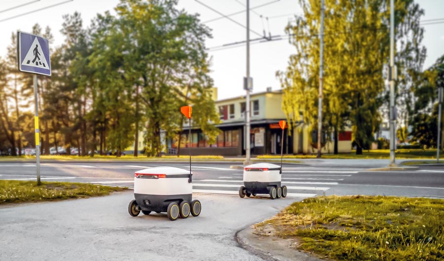 Two cooler-sized robots on the sidewalk, headed toward a crosswalk.