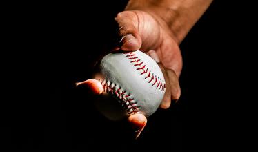 A hand holds a baseball against a black background. The wrist is showing and the thumb covers the top of the ball, with the palm behind the ball and the fingers curling underneath.