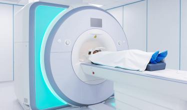 A person is on their back, covered in a white cloth, at the entrance to an MRI machine, with blue coverings on their feet. The MRI machine is mostly white, with an aqua-colored glow along one side.